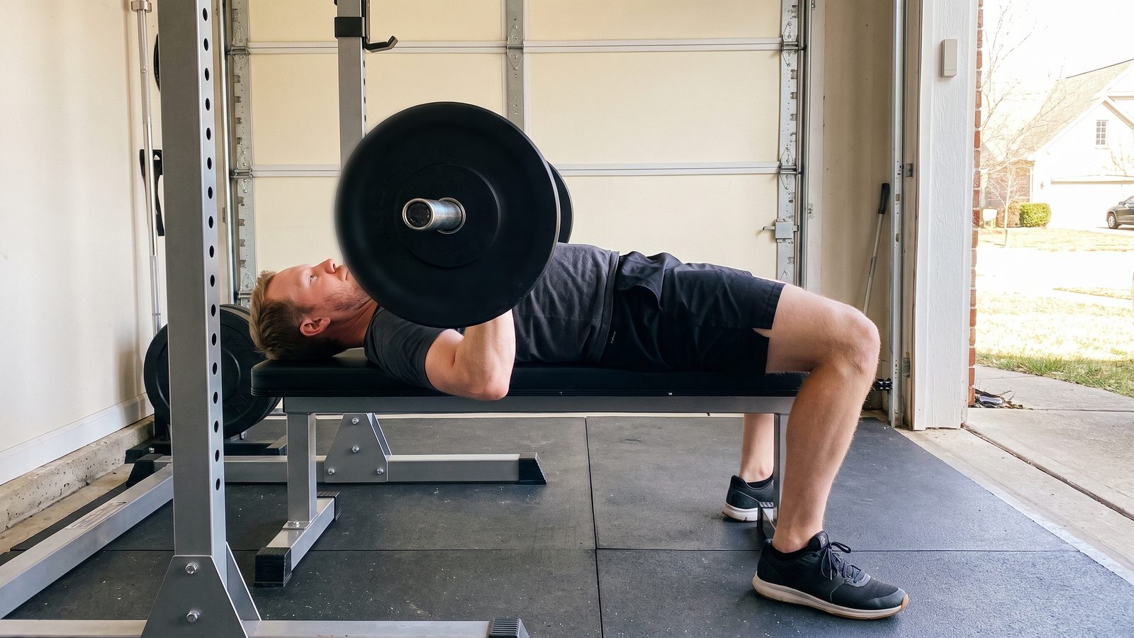 A man at the bottom of a barbell bench press in a garage gym, seen from the side at bench height, the loaded bar touching his chest and his feet flat on the floor