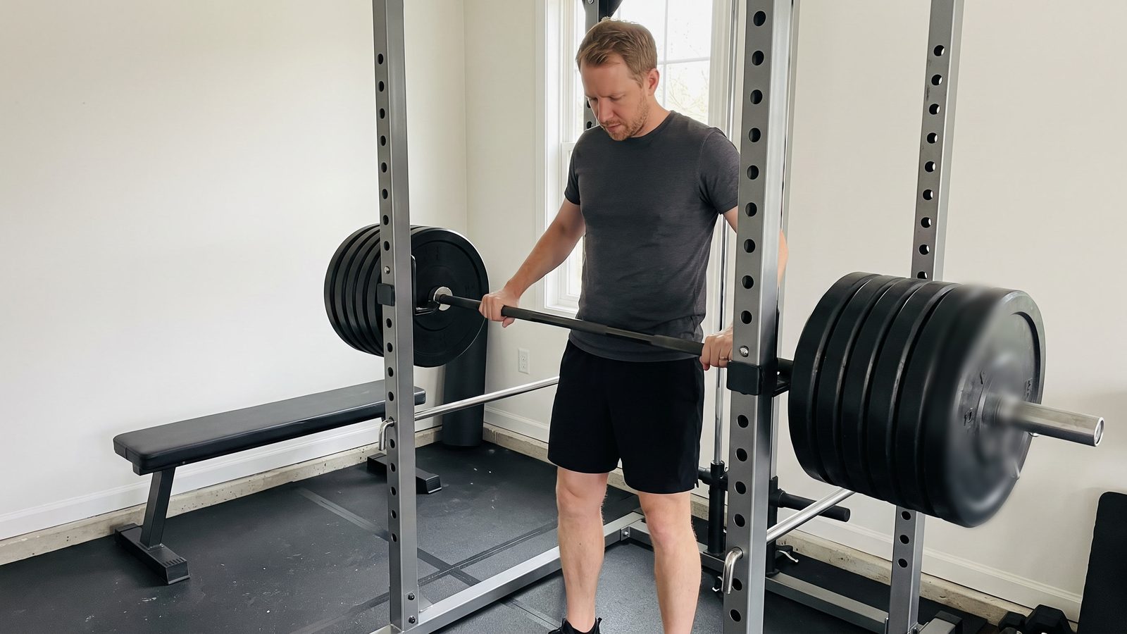A man standing inside a power rack in a garage gym with one hand on a heavily loaded barbell resting in the hooks, looking down at it before a set