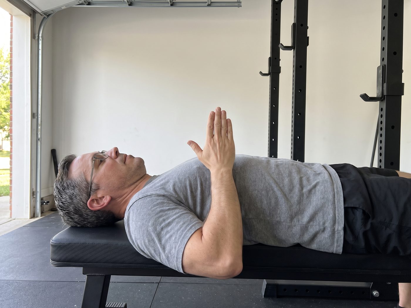 A man in glasses lying on a flat bench in a garage gym with no bar, holding one hand edge-on and upright on his chest to show the height where his press stalls