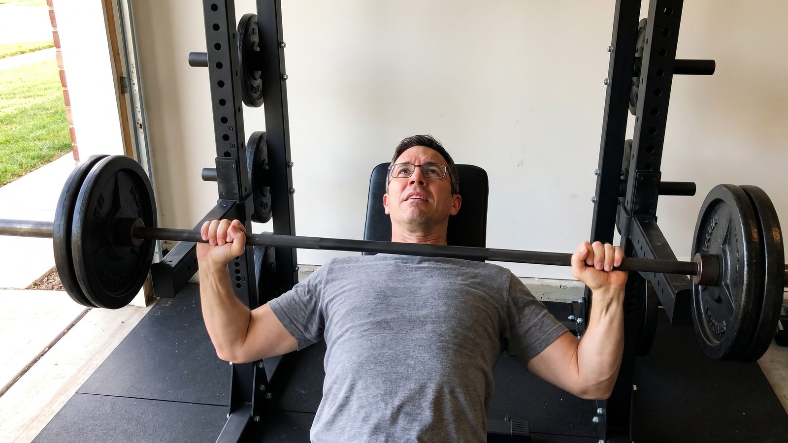 A man in glasses on a flat bench in a garage gym, a straight barbell loaded with two plates a side stalled a hand above his chest, photographed from the foot of the bench