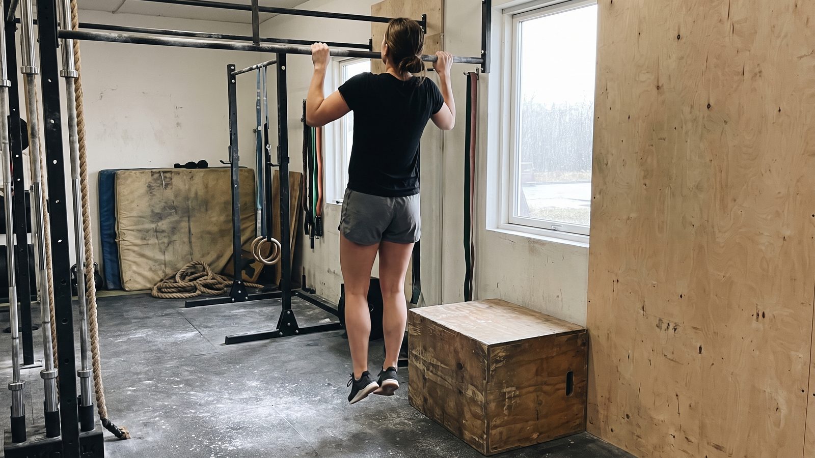 A woman who has stepped off a wooden box under a pull-up bar, chin at the bar and feet hanging free, beginning a slow descent