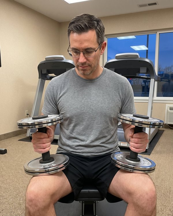A man in glasses seated on a hotel gym bench holding two plate loaded dumbbell handles