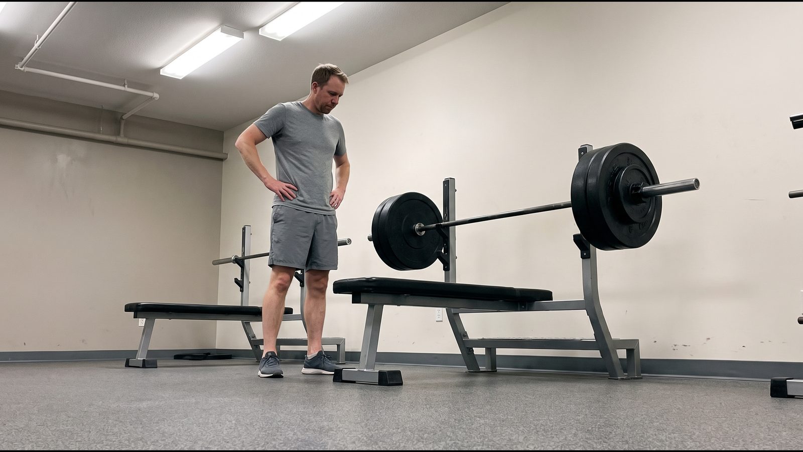 Two flat benches side by side in a quiet gym, a lifter standing beside one of them looking down at a loaded barbell
