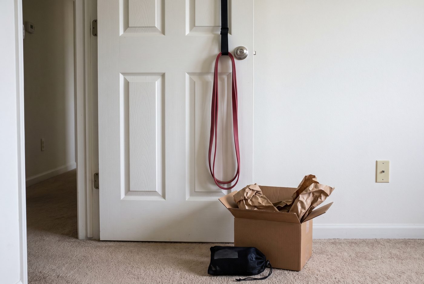 A single red resistance band hanging from a door anchor at the top of a bedroom door, with an opened shipping box on the carpet below