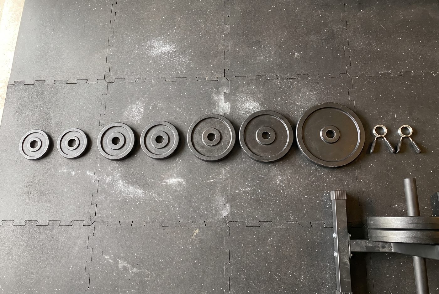 Small iron weight plates laid out in a row on a garage floor from 2.5 pounds up to 25, next to two spring collars
