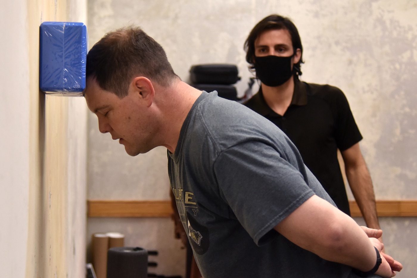 A man presses the side of his head into a foam block on a wall while a trainer stands behind him
