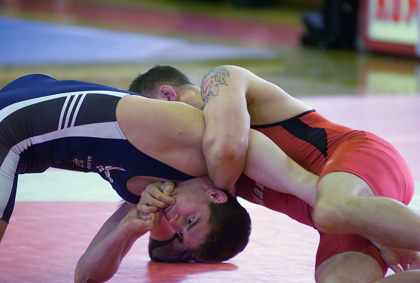Two wrestlers on a red mat, the one underneath driven onto the crown of his head under his opponent's weight