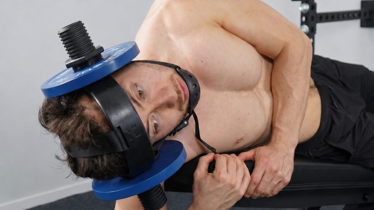 A man lying back on a bench doing a neck extension in a Kensui EZ-HELM loaded with blue weight plates