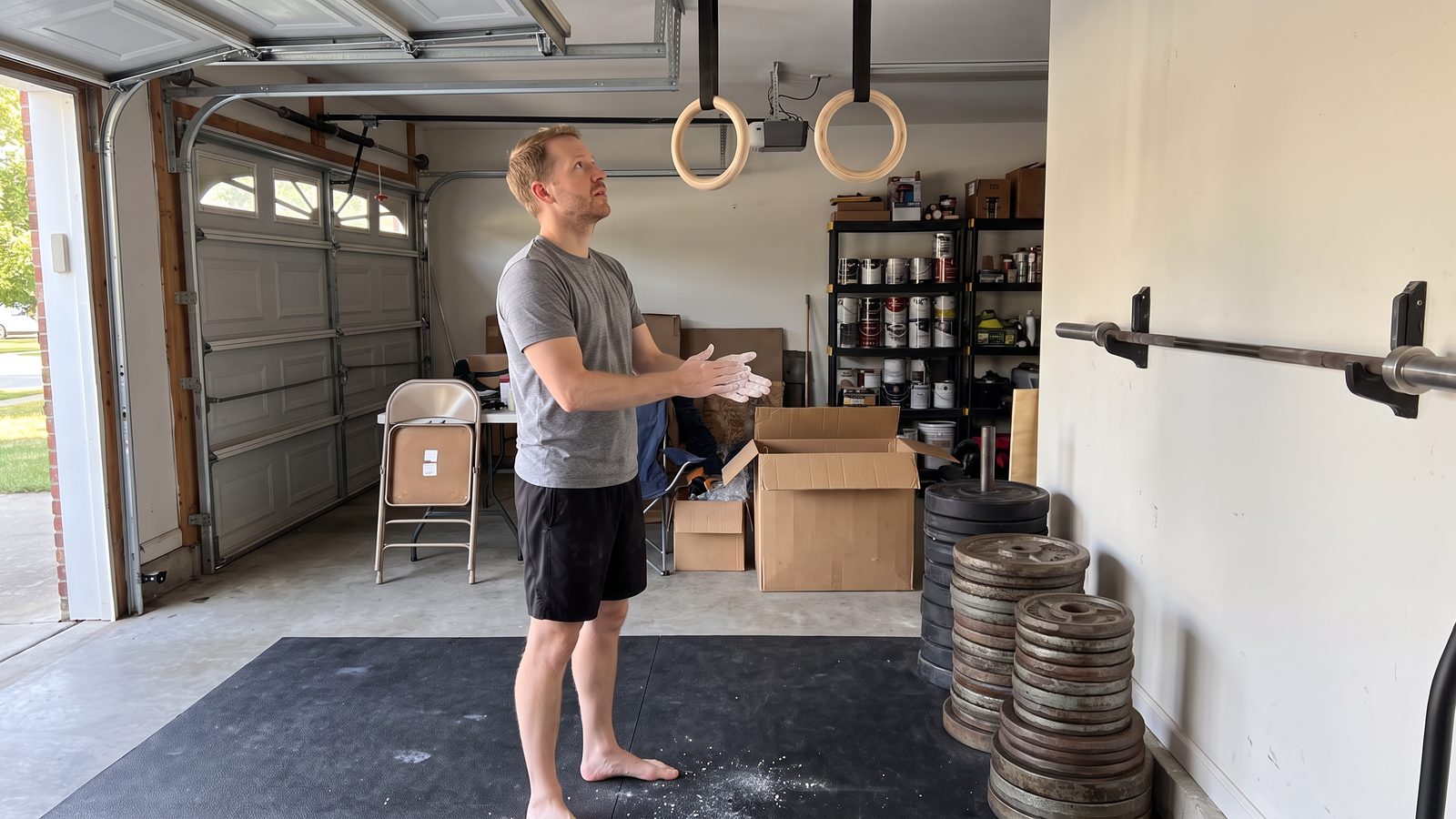 A man chalking his hands under wooden gymnastic rings in a home garage, with stacks of old grey iron weight plates against the wall