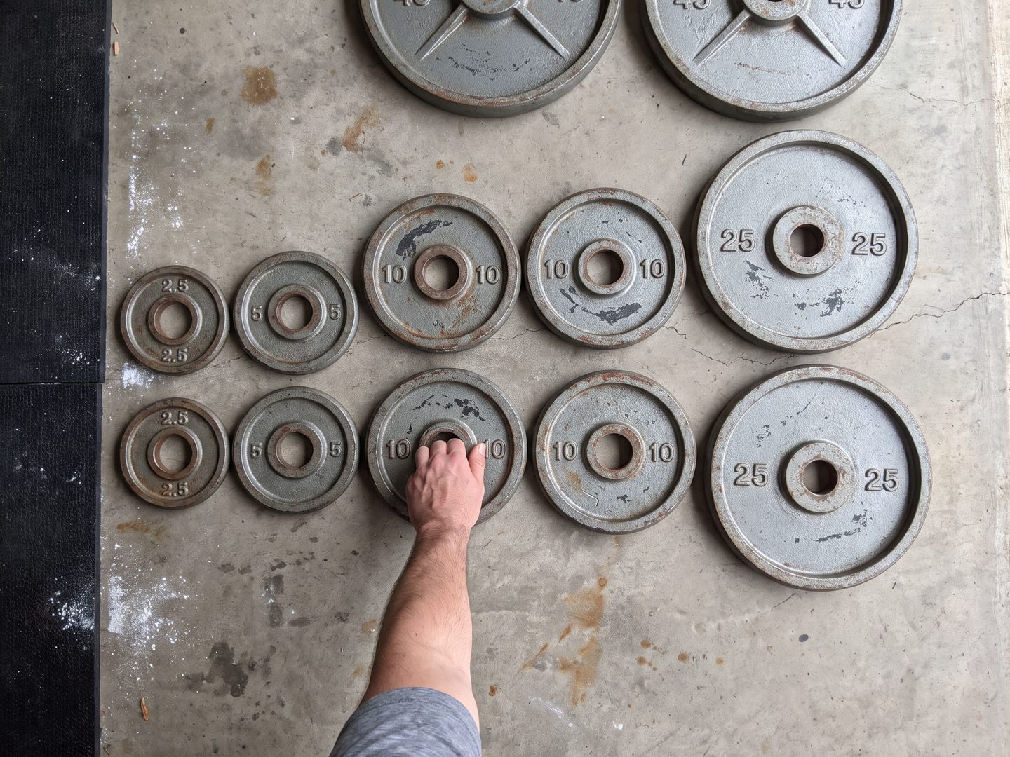 Old grey iron weight plates laid out on a garage floor from 2.5 pounds up to 45 pounds, with a hand reaching for a 10 pound plate