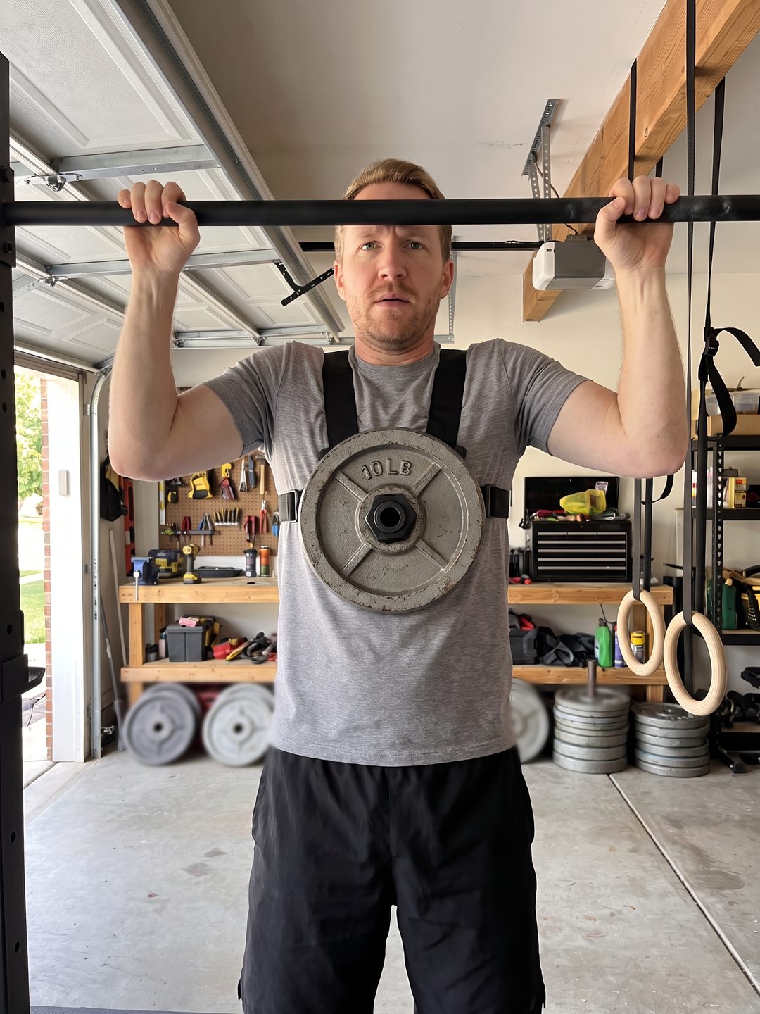 The writer doing a weighted pull-up in his garage wearing a black plate-loaded vest with one of his old 10 pound plates on the chest