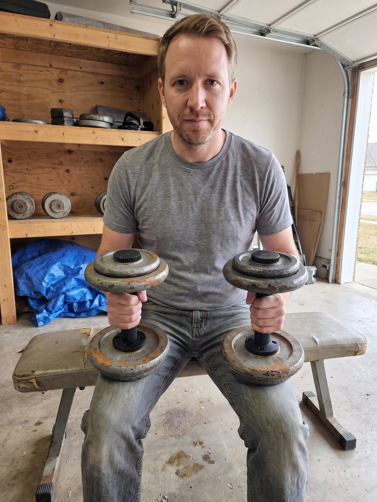 The writer seated on an old weight bench in a garage with two loaded dumbbell handles standing flat on his thighs