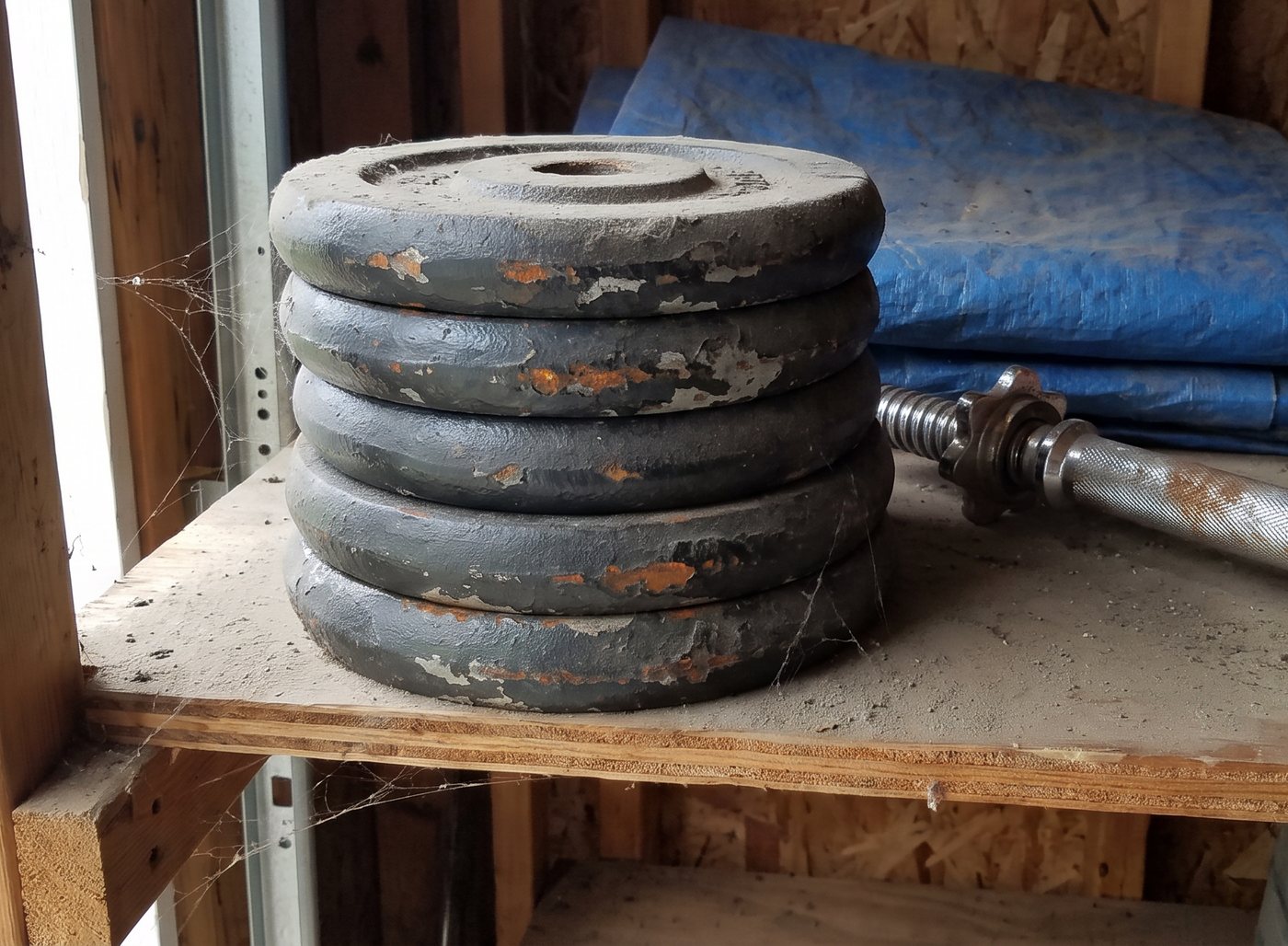 A stack of old grey cast iron weight plates coated in dust on a garage shelf, next to a chrome dumbbell bar and a folded blue tarp