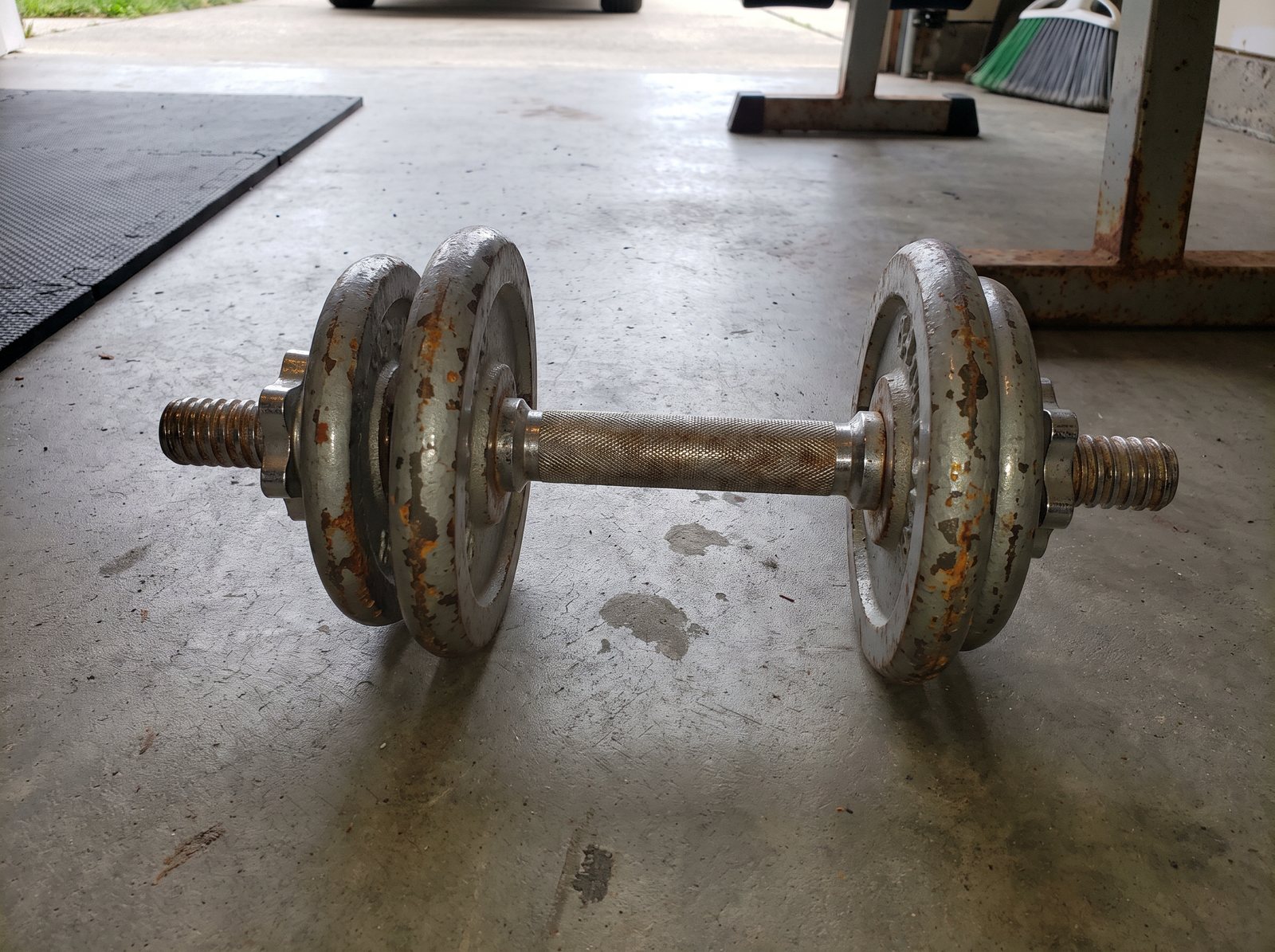 An old chrome spinlock dumbbell on a garage floor, two rusty grey plates on each end and bare threaded bar sticking out past both star collars