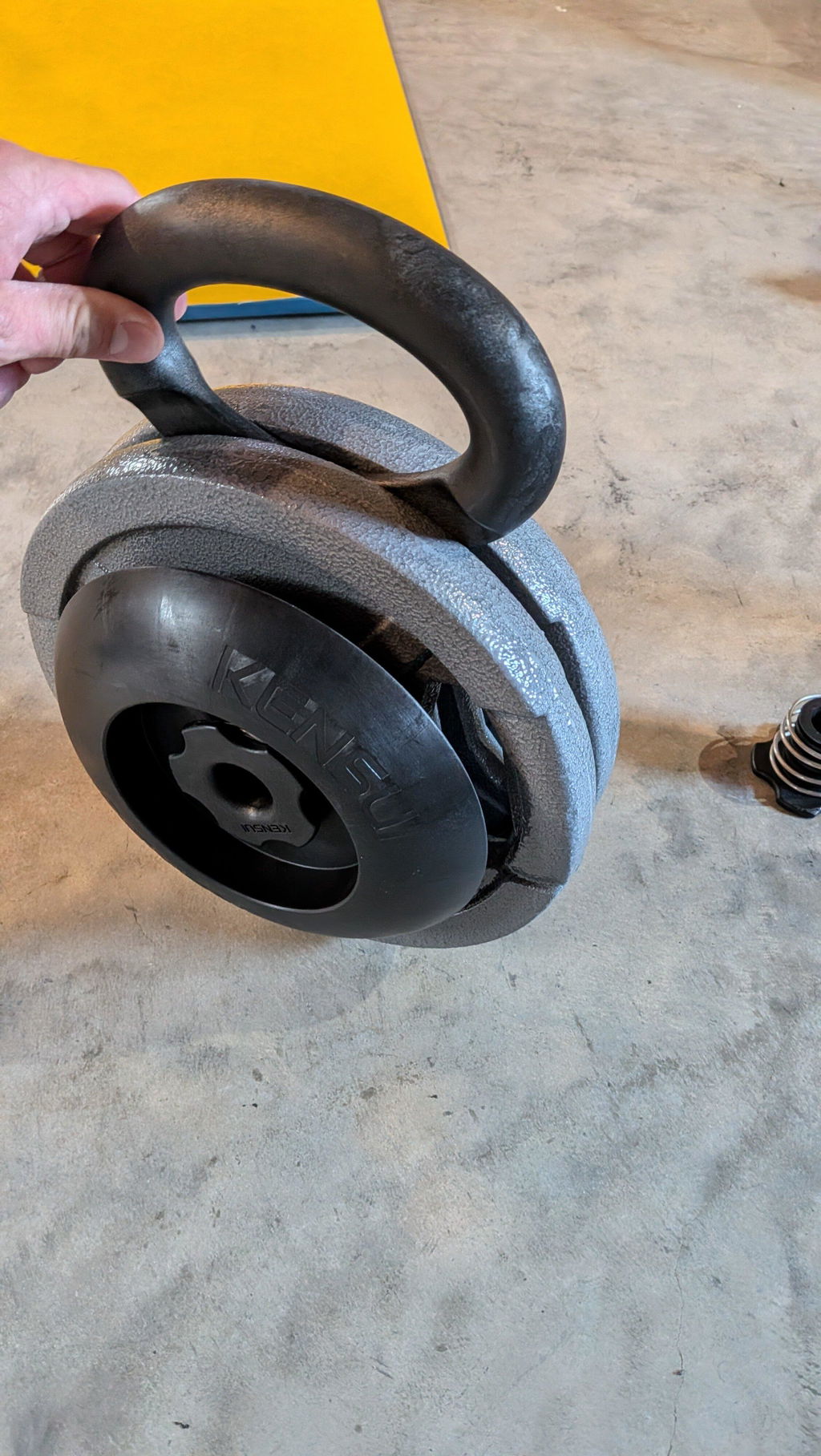 A hand lifting a Kensui EZ-BELL loaded with grey iron plates off a gym floor