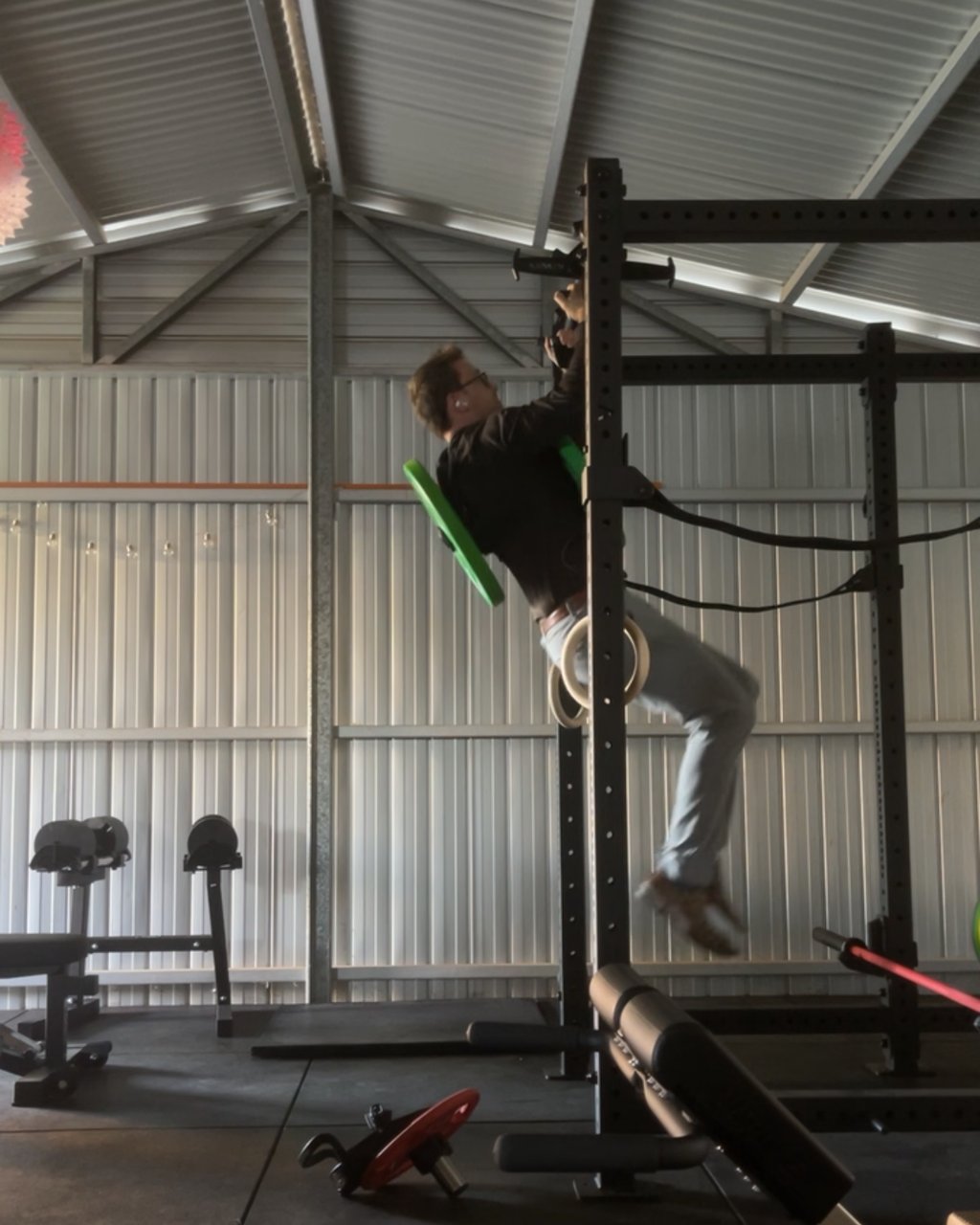 A man doing a weighted pull-up on black neutral grip handles hung from the bar of a power rack in a metal shed gym