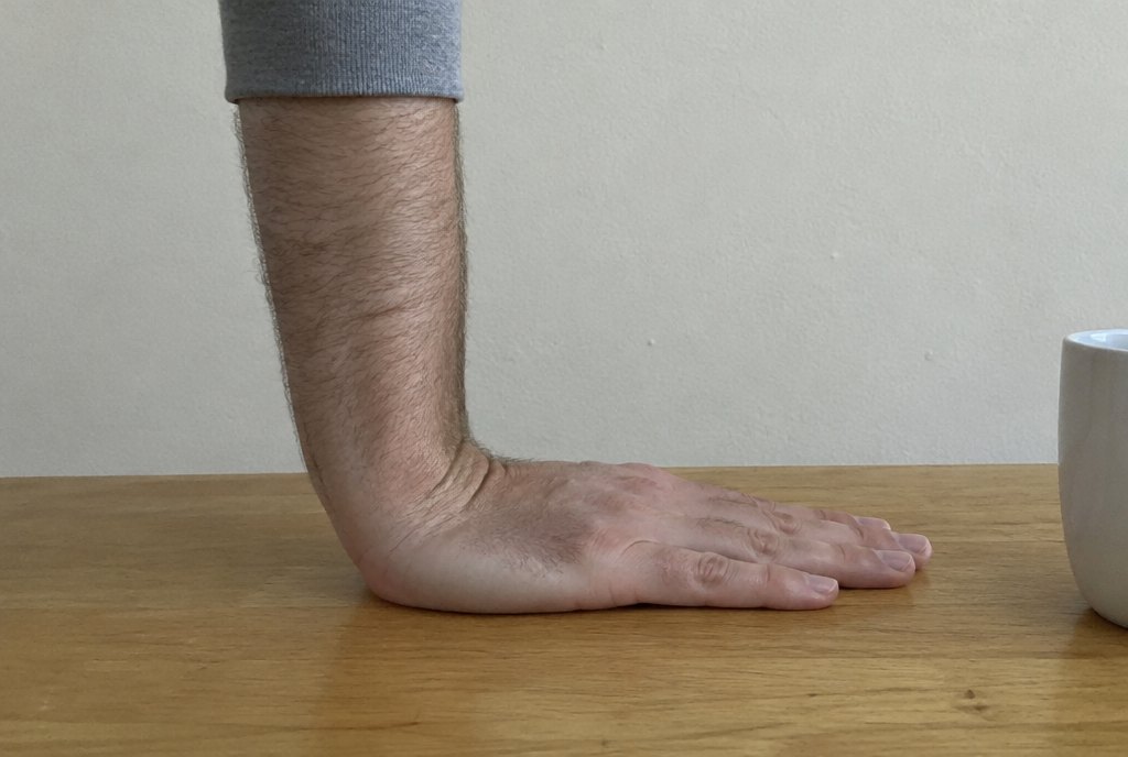 A hand pressed flat on a wooden table with the forearm standing straight up from it, the wrist at a right angle