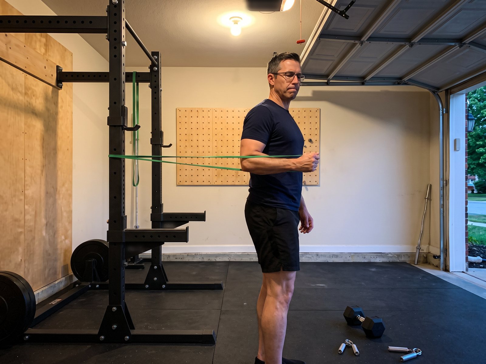 The author in his garage doing a shoulder rotation with a green resistance band looped around the rack, a light dumbbell and two hand grippers on the floor