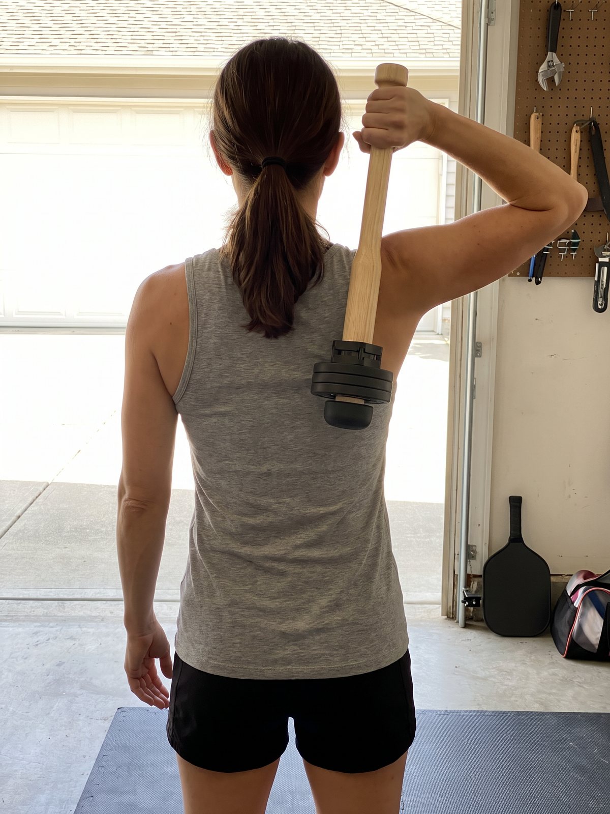 The author's wife, seen from behind in the garage with the door open, holding a wooden club with a small plate on the end so it hangs behind her right shoulder, a paddle by the door