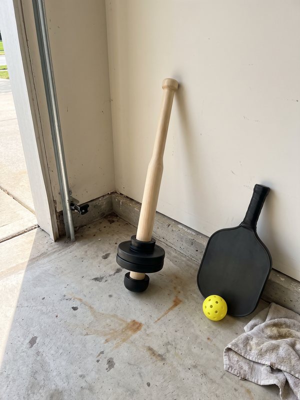 The wooden EZ-CLUB PRO standing on two small black plates in a garage corner next to a pickleball paddle