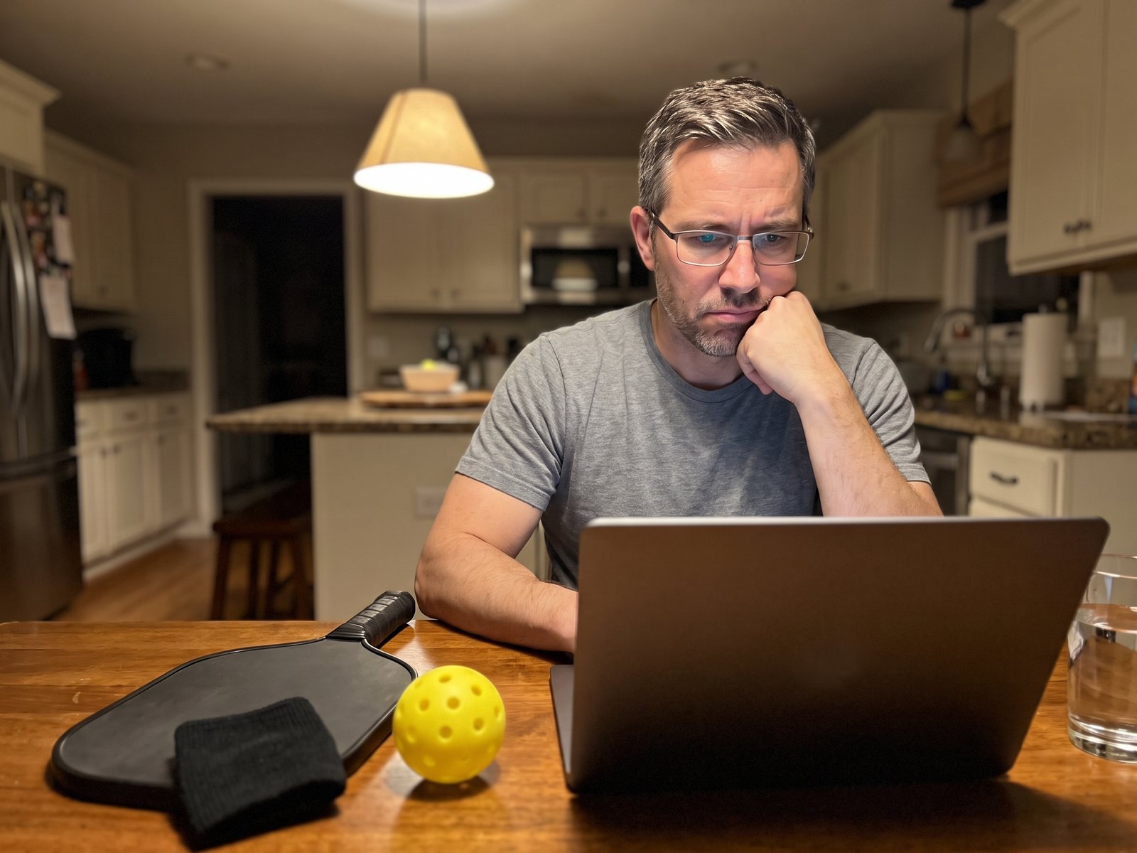 The author at the kitchen table in the evening, chin on his fist, reading a laptop with his paddle and wrist sleeve beside it