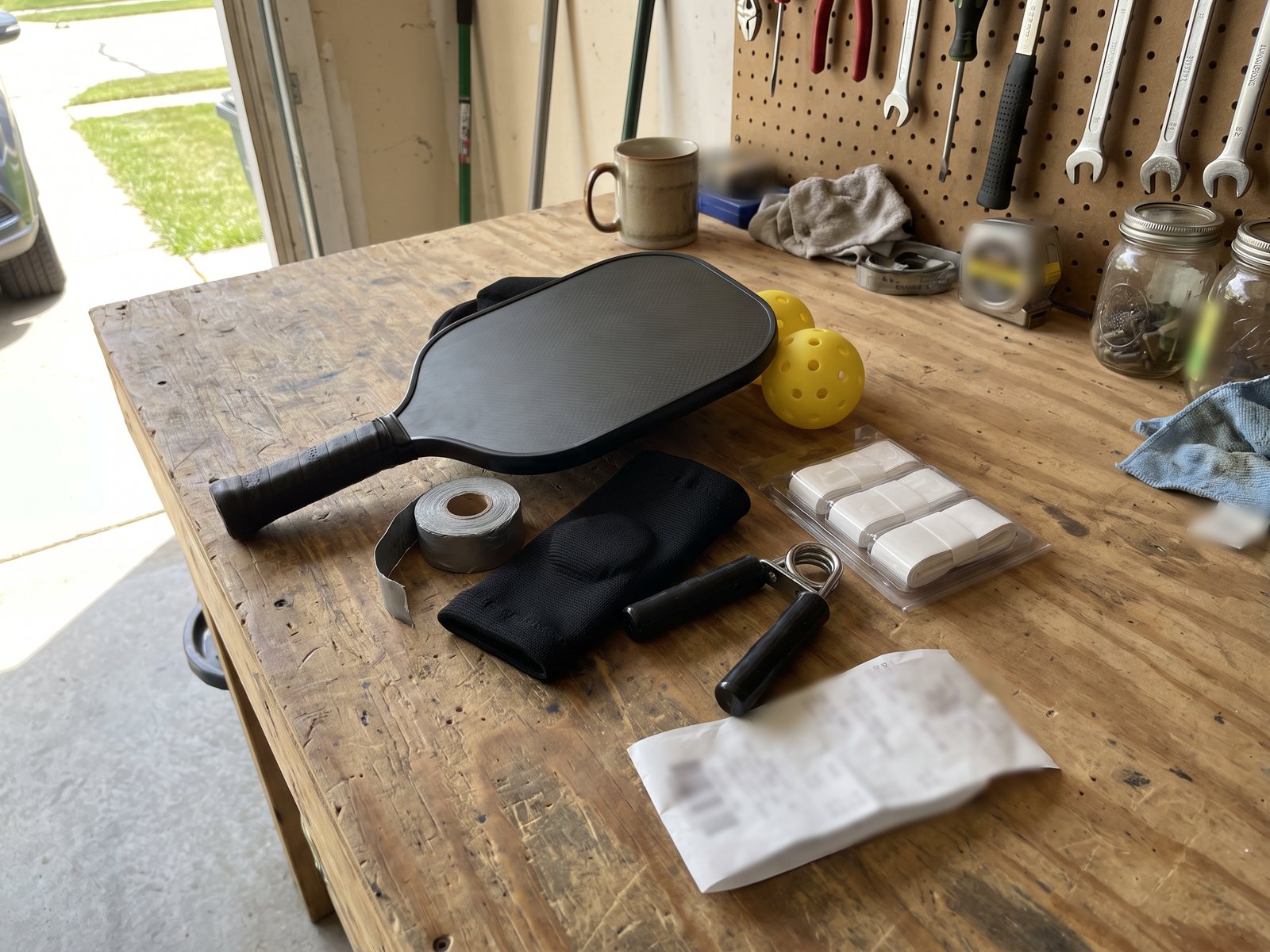 A new paddle, a roll of lead tape, a black wrist sleeve, a spring hand gripper, overgrips and two pickleballs laid out on a garage workbench