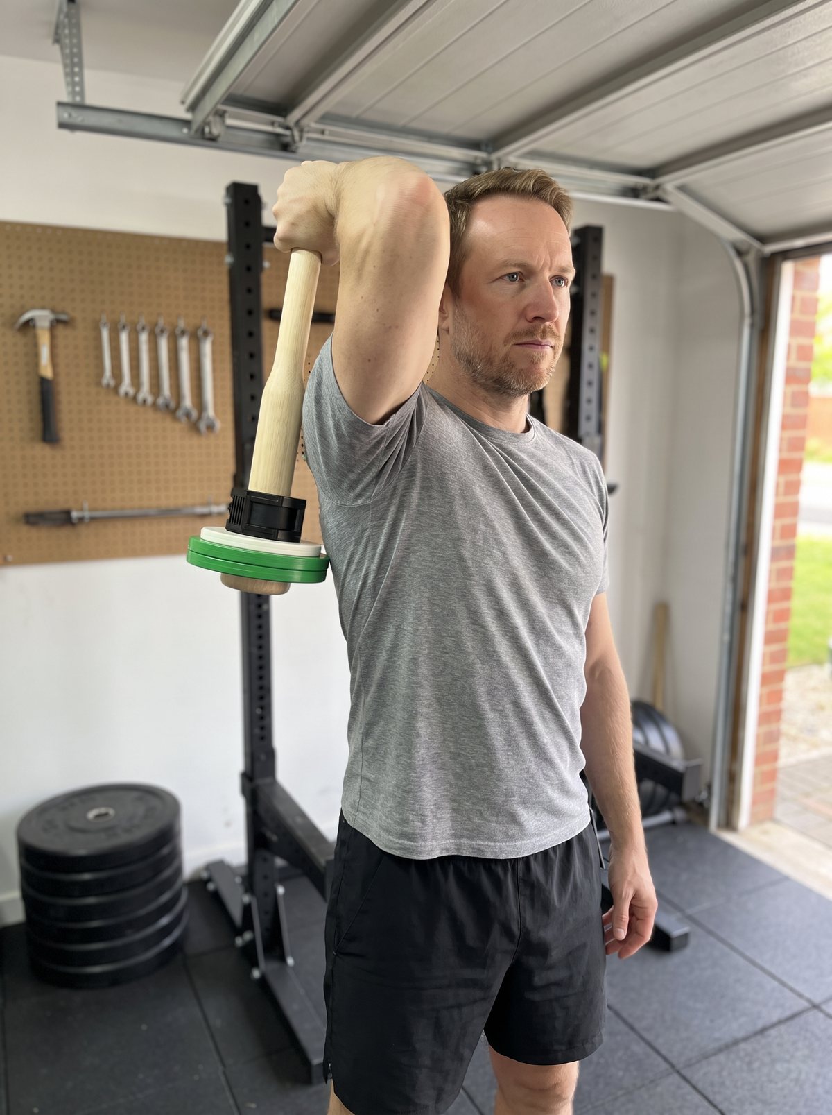 The author in his garage holding the wooden club behind his head with one hand, elbow pointing up, plates hanging below
