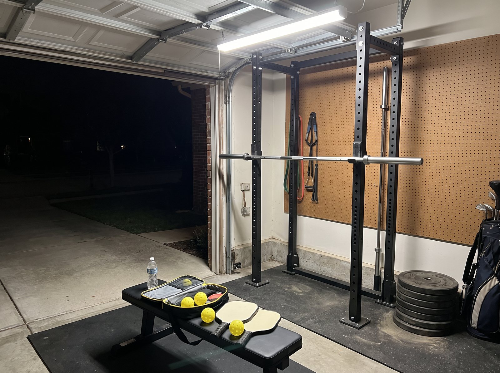 The author's garage gym at night, a pickleball bag and paddles lying on the weight bench in front of an empty barbell
