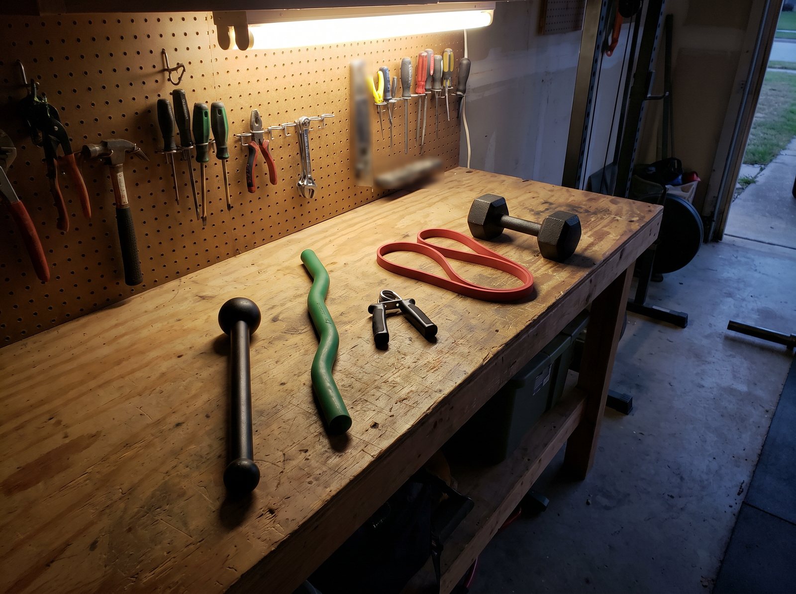 A small fixed steel club, a rubber twisting bar, a hand gripper, a resistance band and a small dumbbell on a garage workbench