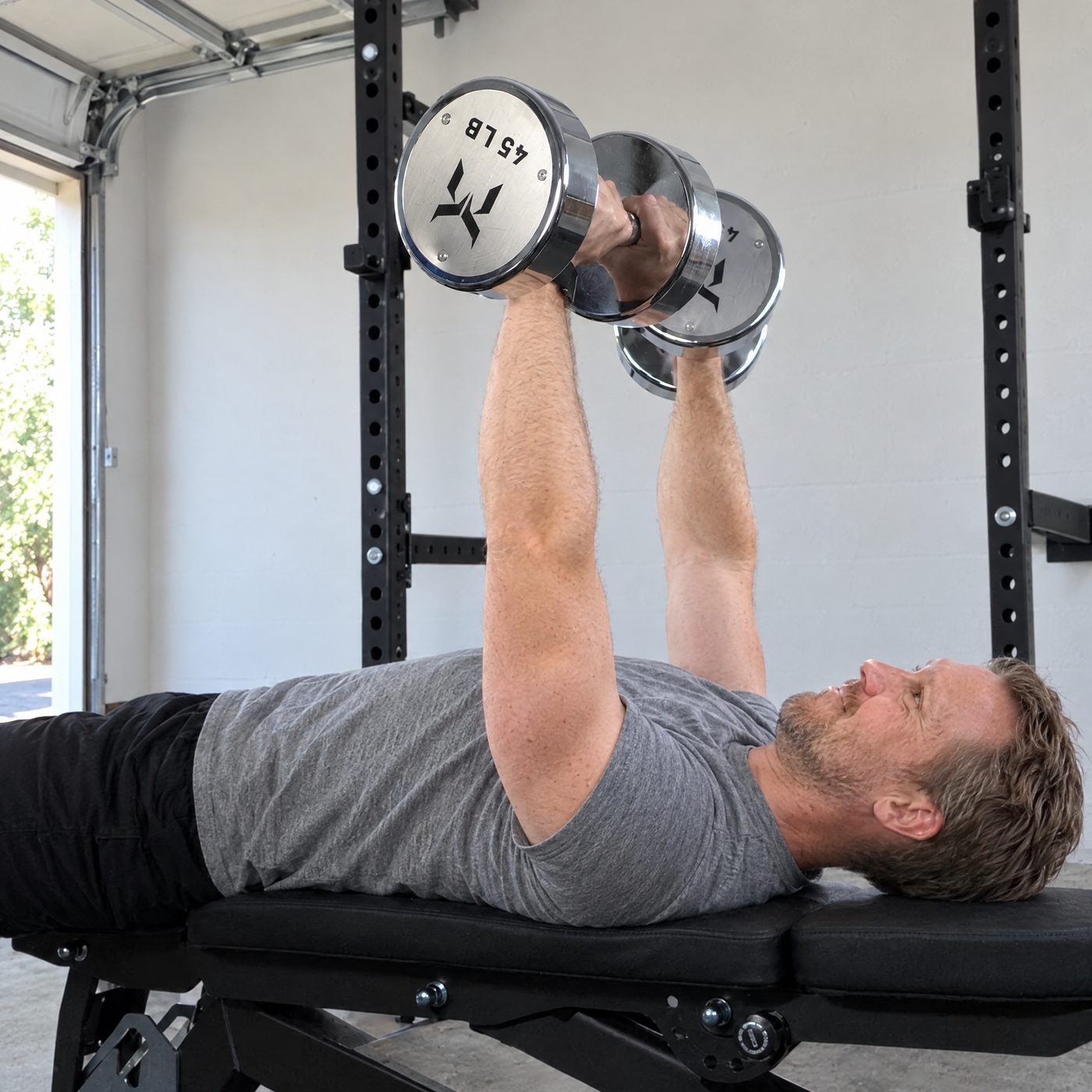 A man lying on a flat bench in a garage gym pressing two chrome 45 pound dumbbells
