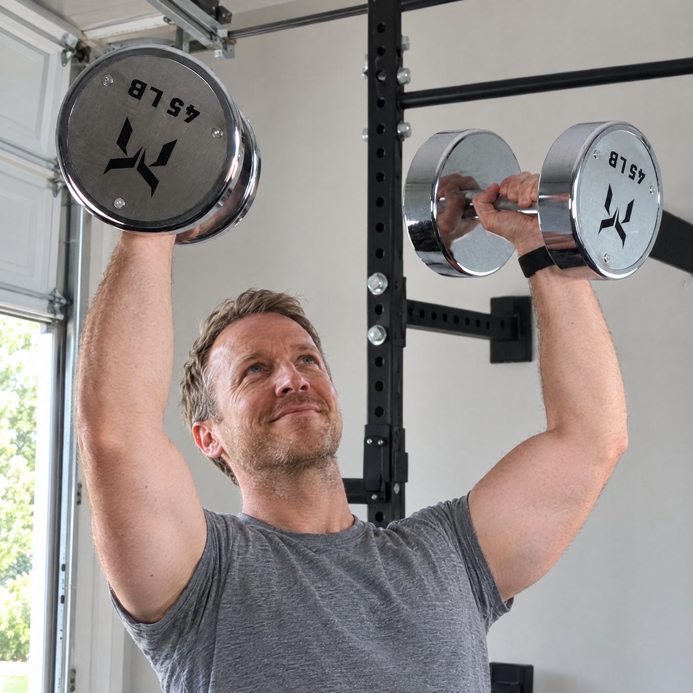 A man in a grey shirt pressing two chrome 45 pound dumbbells overhead in a garage gym