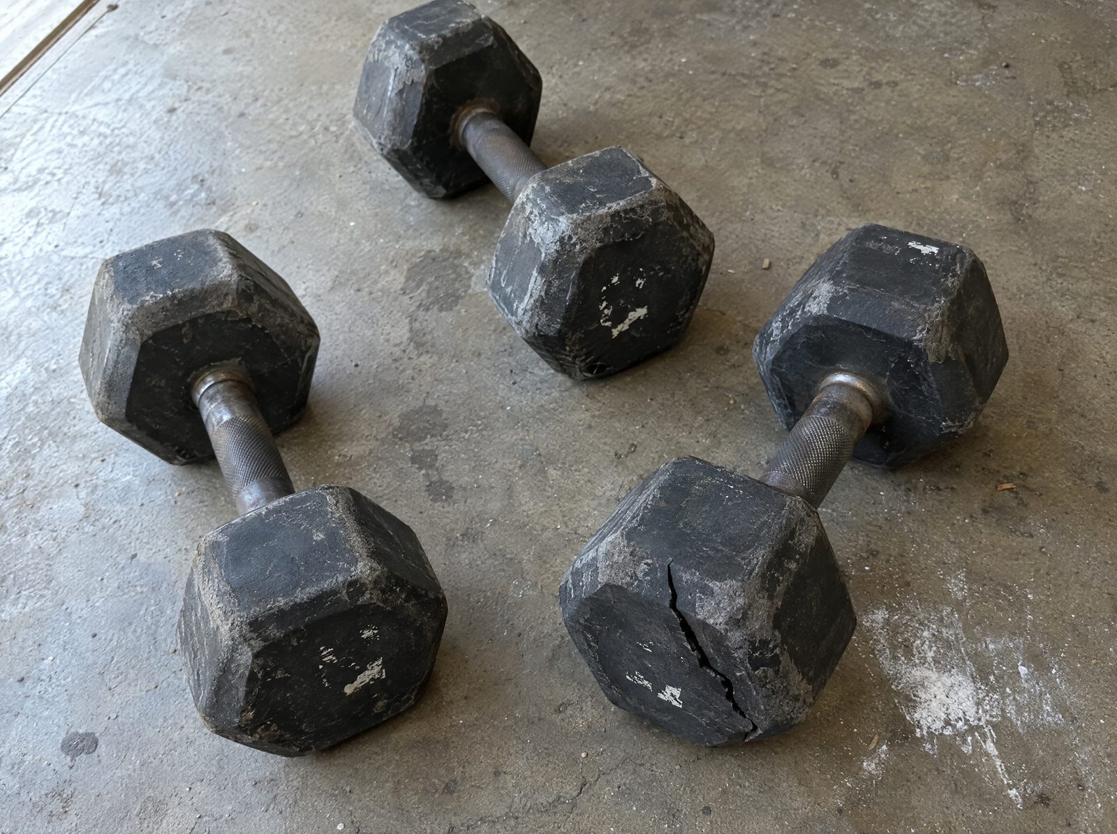 Three scuffed rubber hex dumbbells on a concrete floor, one head cracked across, the painted numbers almost gone