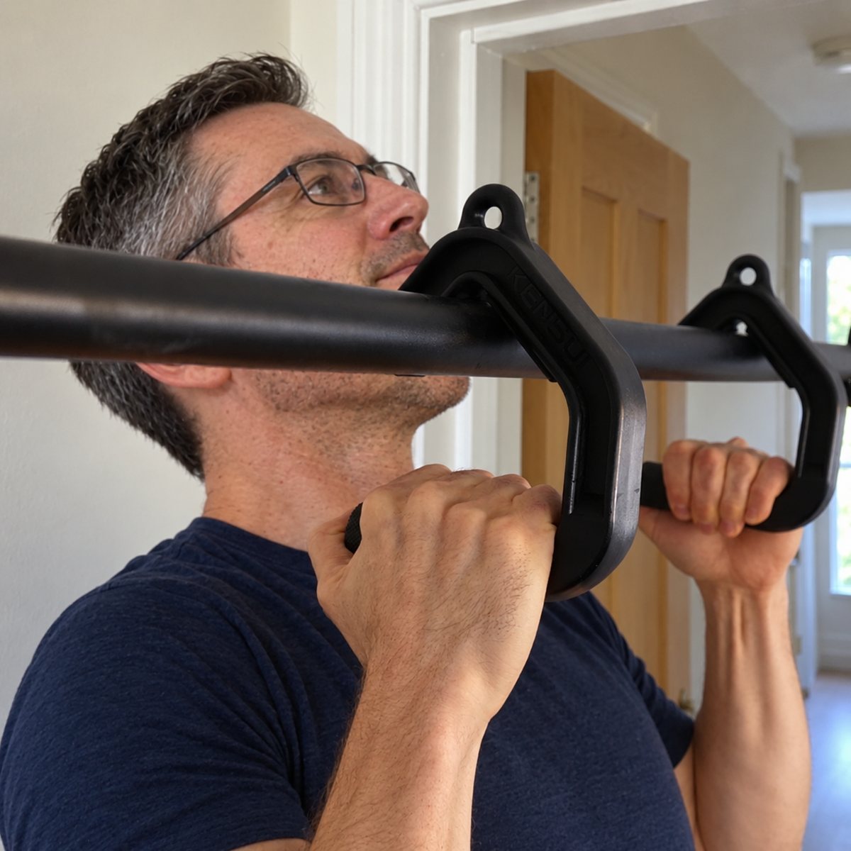 A man in glasses and a navy t-shirt doing a pull-up on black handles hung from a doorway bar, palms facing each other