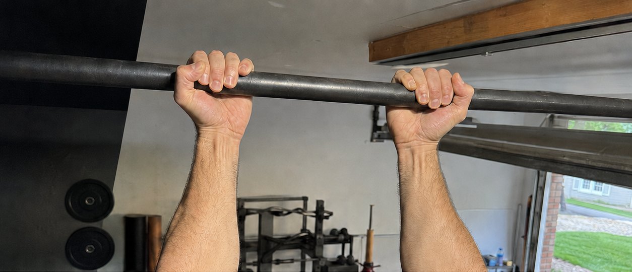 Two bare hands gripping a plain steel pull-up bar overhead, palms facing forward, in a home garage
