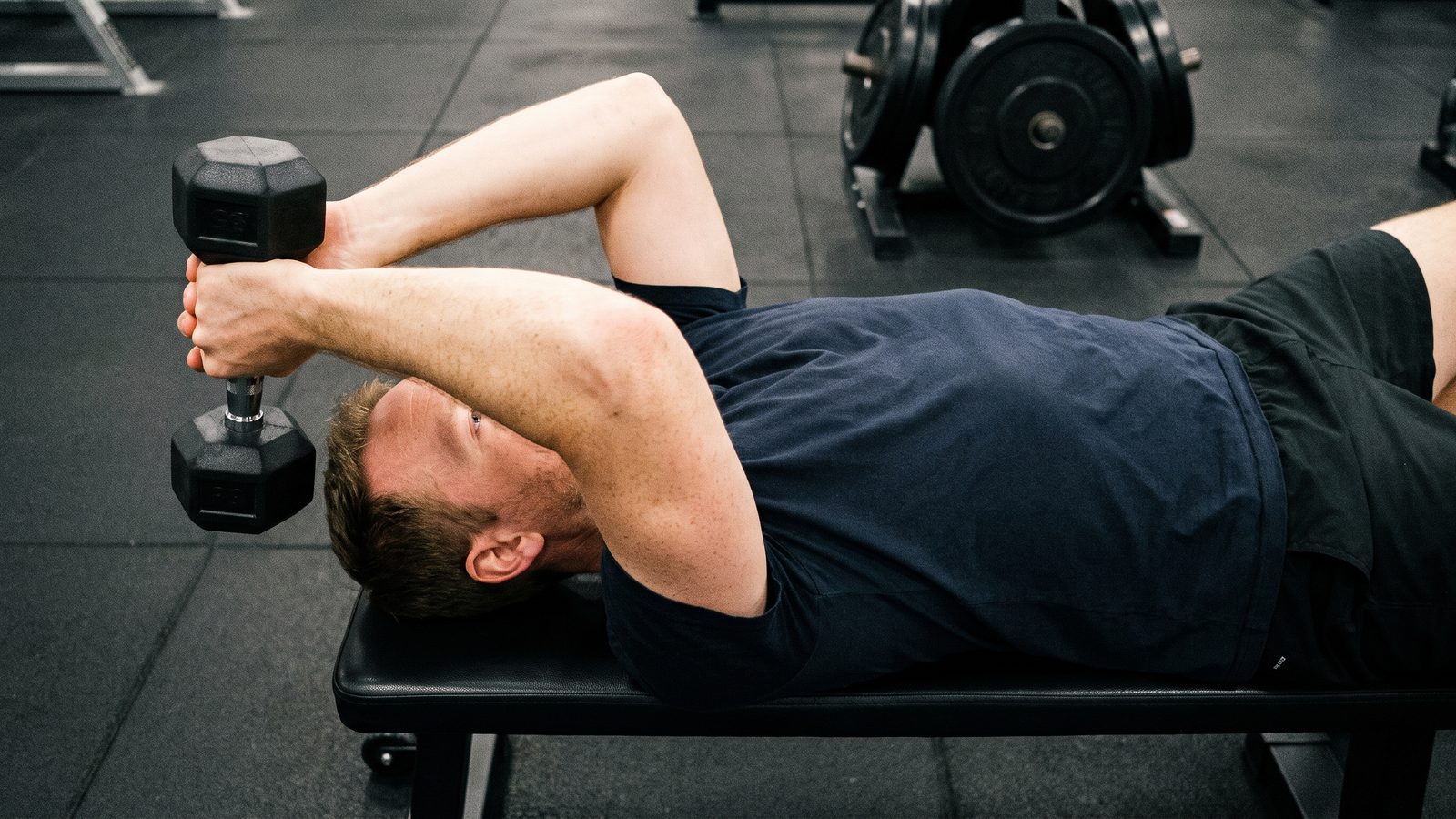 A man in a navy t-shirt lying on a flat bench in a gym, holding a black hex dumbbell in both hands behind his head with his elbows pointing up, at the bottom of a lying triceps extension, a plate tree out of focus behind him