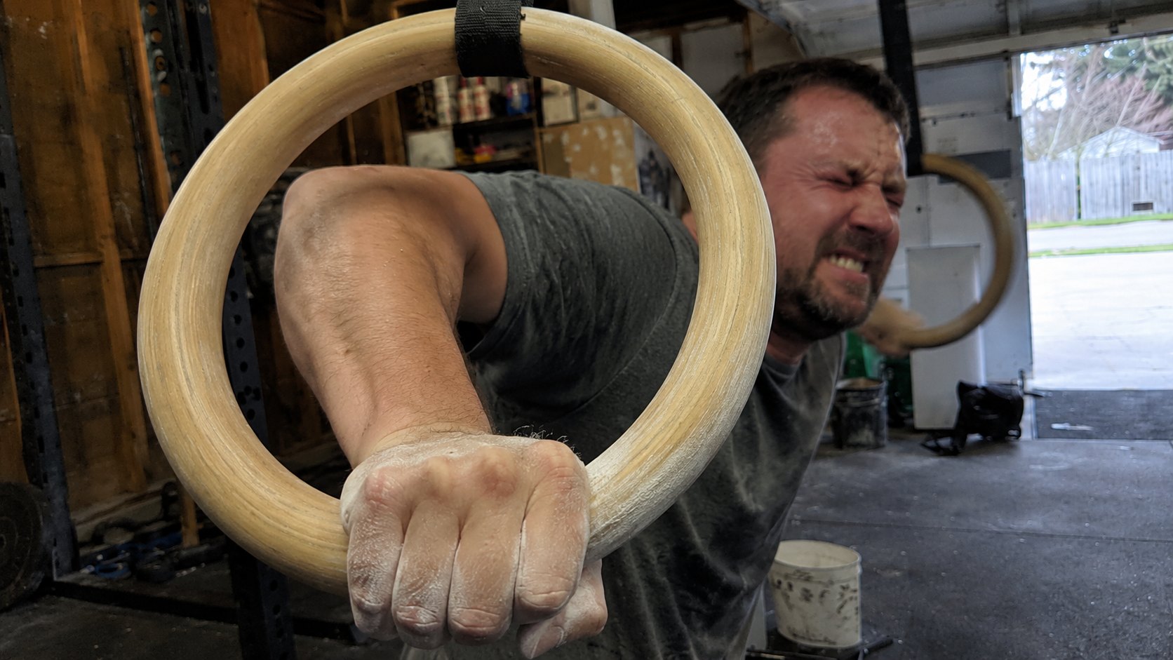 A man grimacing with effort in a garage gym, one chalked hand gripping the bottom of a plain wooden gymnastic ring that fills the frame