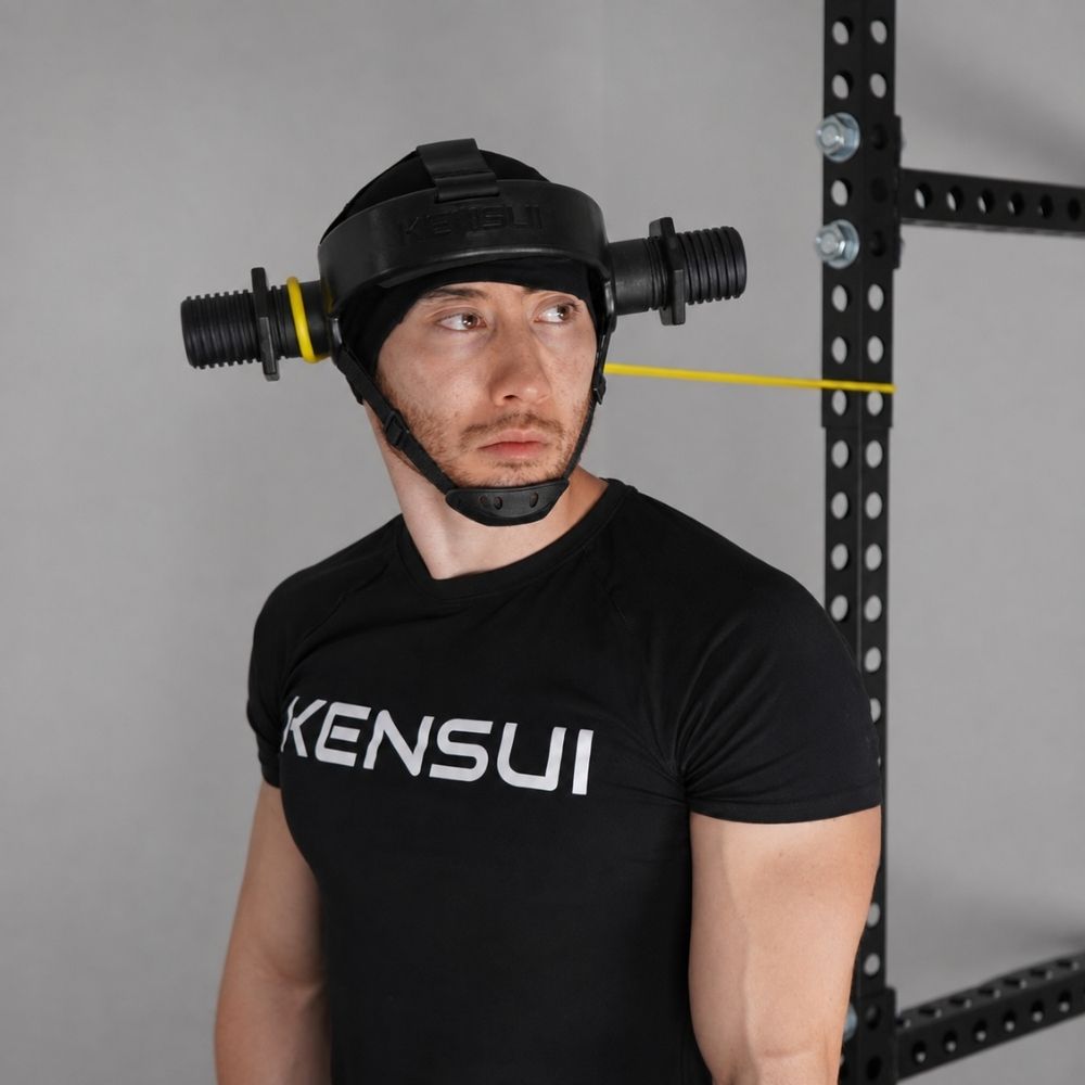 A man in a black t-shirt wearing the plate-loaded neck helmet with a yellow resistance band clipped to the ear sleeve, slowly turning his head against the band in a squat rack