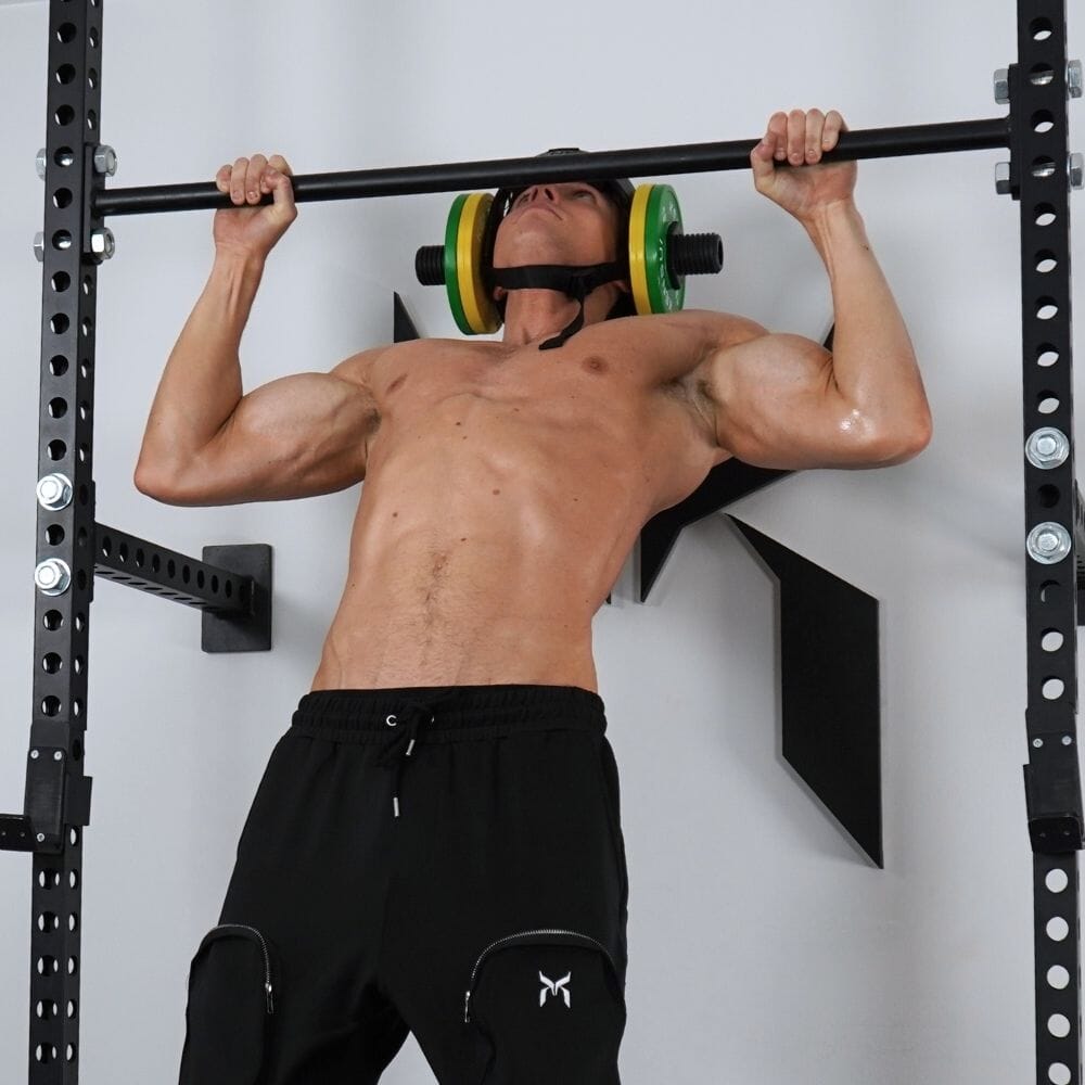 A shirtless man hanging from a pull-up bar in a gym with the loaded neck helmet on, a green and a yellow plate at each ear, holding his head level