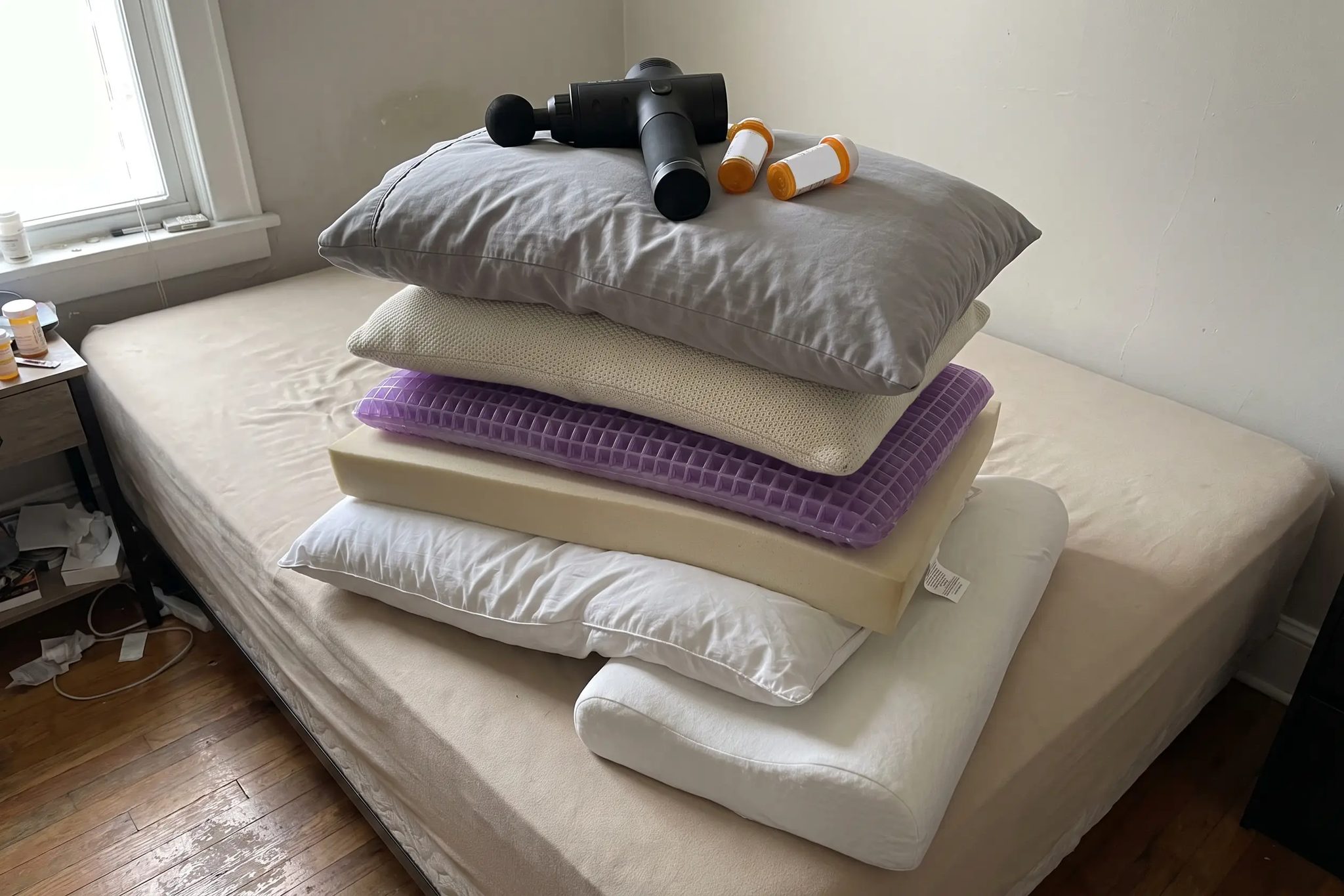 Six different pillows stacked on a bare mattress in daylight, a black massage gun resting on top and two orange prescription bottles standing beside the pile
