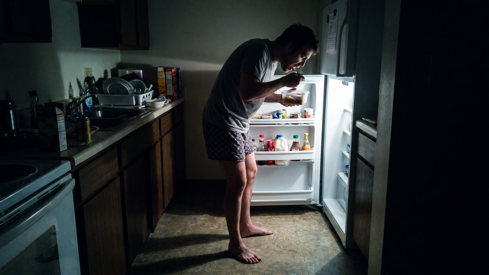 A man eating from a container in front of an open refrigerator in a dark kitchen at night